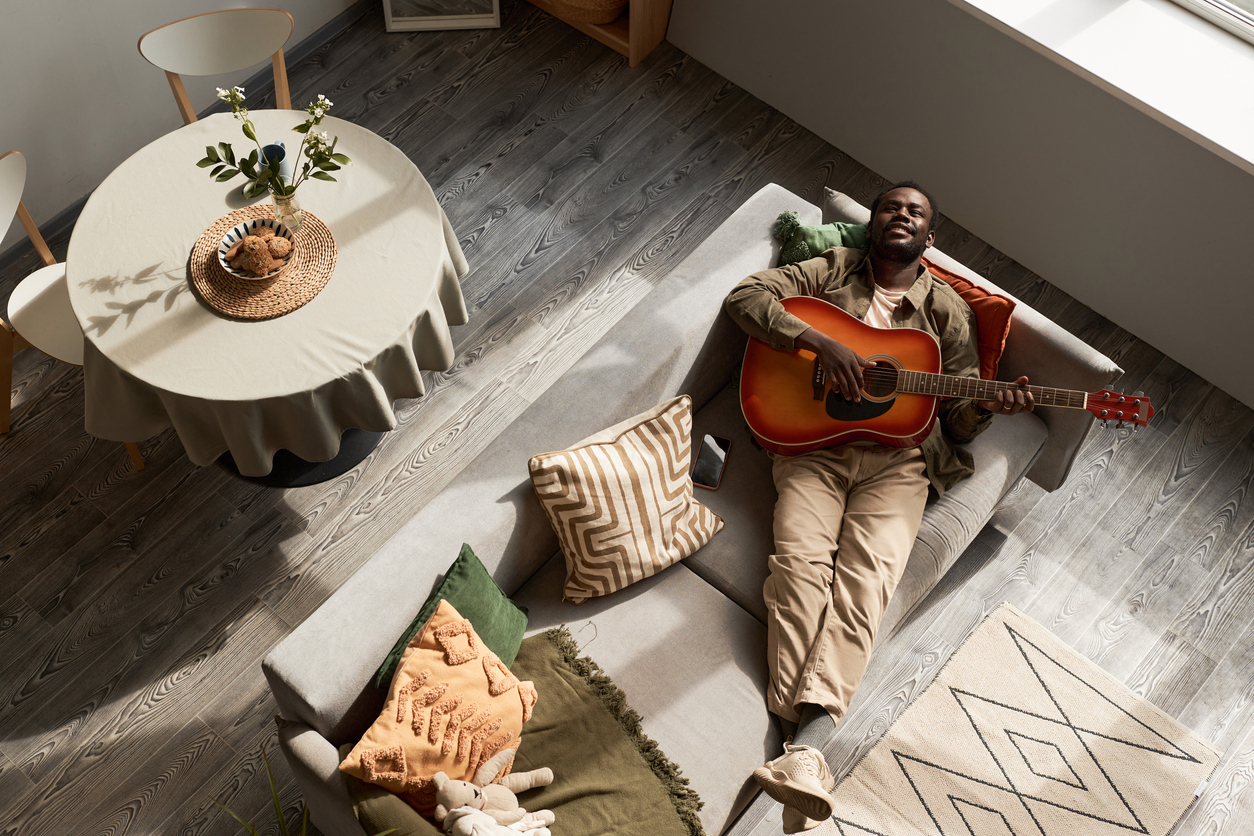 Top down view of Black young man playing acoustic guitar lying on sofa in cozy home lit by sunlight, copy space